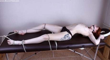 Thin redhead sports a ball gag while tied to a table with rope in her panties