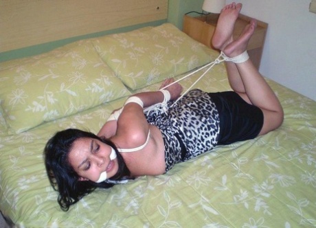 On a bed, the girl with dark hair is covered in her hands and tied loosely while wearing a dress.