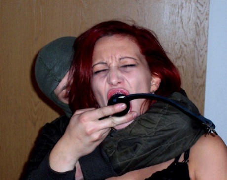 Lilu Natilova, a redhead with no hair visible, is tied up and gagged by an individual wearing a hood.