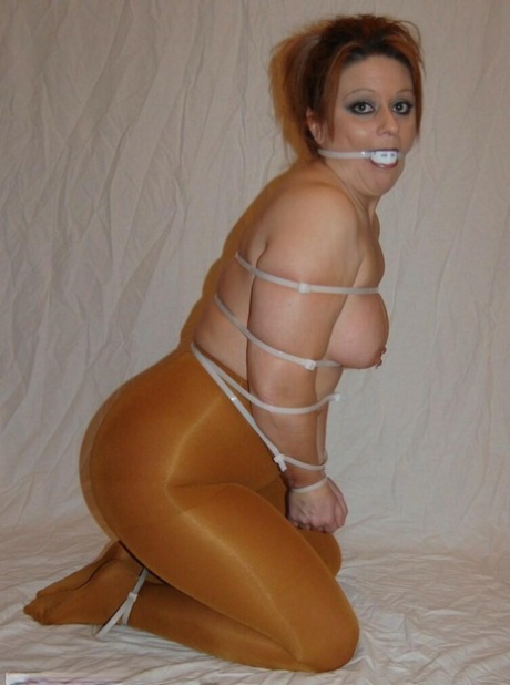 The topless redhead, Adrienna, is being ball gagged while being wrapped in hose.