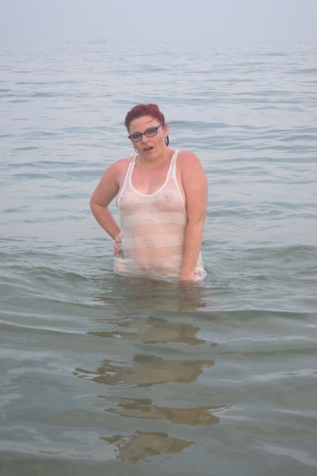 An inked reddish woman wades through the water and exposes her tattoo before venturing into the sea.