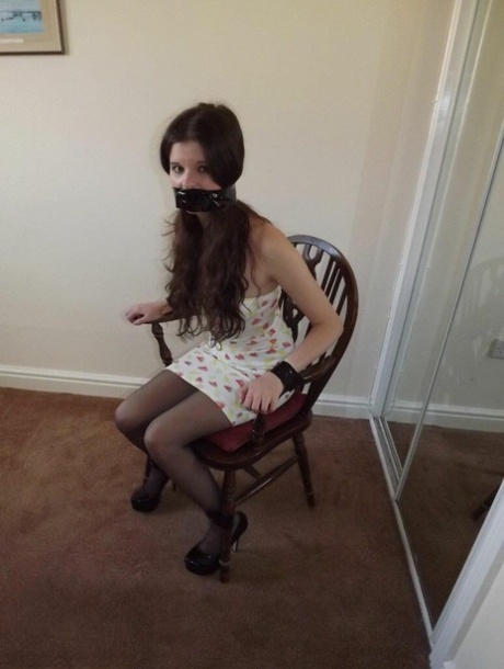 The girl Tamara, dressed in clothes, is unable to move and appears to be gagged and restrained in a chair while staring at a mirror.