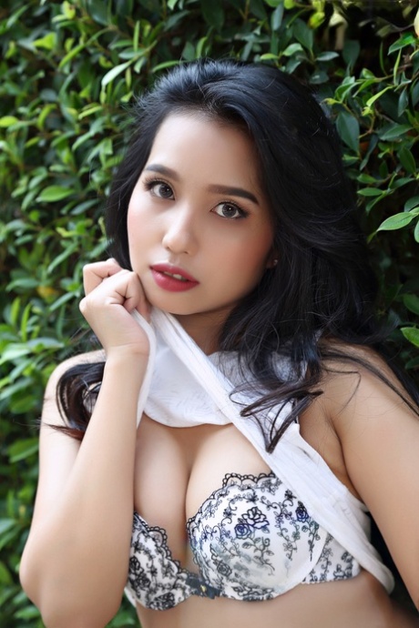 In a garden, Norah, a beautiful Asian girl, is seen in a naked position next to a hedge.