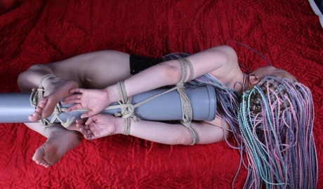 A white girl with bare feet is fastened to a post and placed face down on a bed.