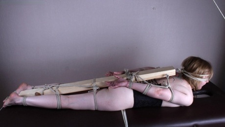 A white woman has her feet, arms and legs fastened to a 2 x 4 that is behind her back.