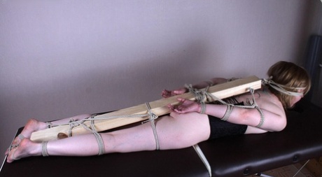 A 2 x 4 is secured behind the back of a Caucasian female, with her feet, arms, and legs connected.