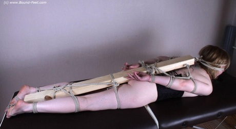 Behind the female figure, a 2'x 4 is being tied to her feet, arms, and legs.