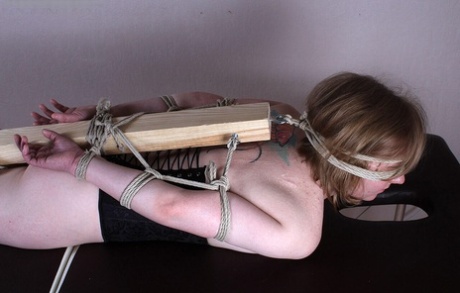 A white woman has her feet, arms and legs fastened to a 2 x 4 that is behind her back.