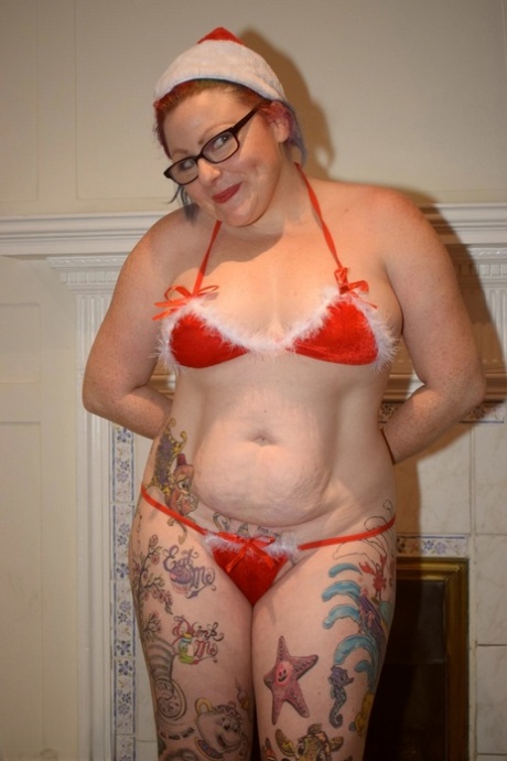 A woman with tattoos gets up from her hat to expose it in a nude bikini pose while wearing a Santa cap.