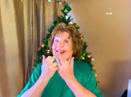 A young Irish plumper named Busty Bliss stands without a top in front of a Christmas tree.