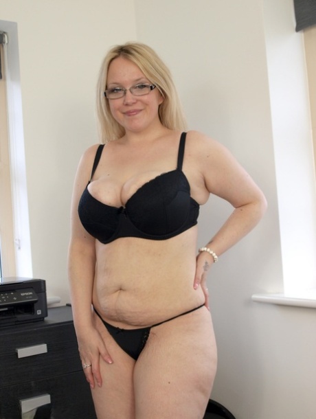 Sindy Bust, a blonde British woman with pale skin, is seen naked on a bed wearing glasses and heels.