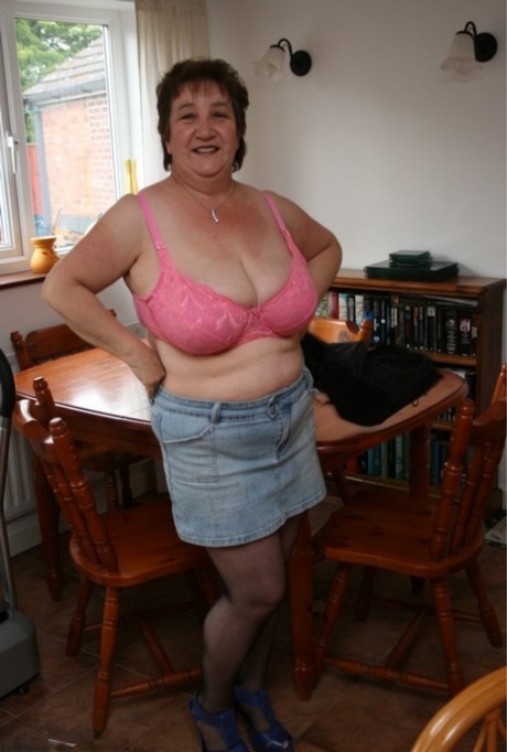 An older British woman named Kinky Carol dresses in a bra and miniskirt while stripping off at a table.