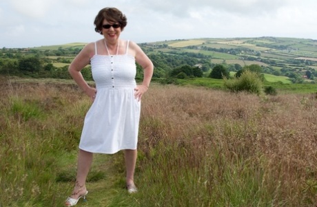 In a field, the elderly lady Dirty Doctor sits on top of a blanket and goes naked.