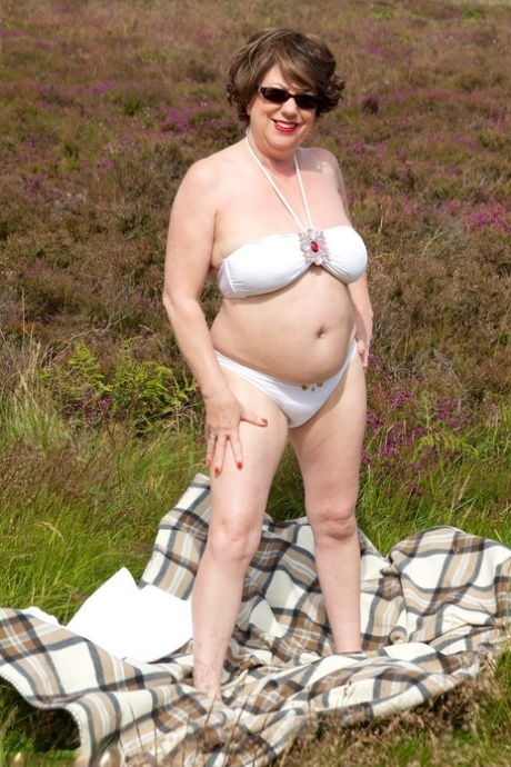 While in a field, the elderly lady Dirty Doctor sits on top of a blanket and enjoys herself without any clothes on.