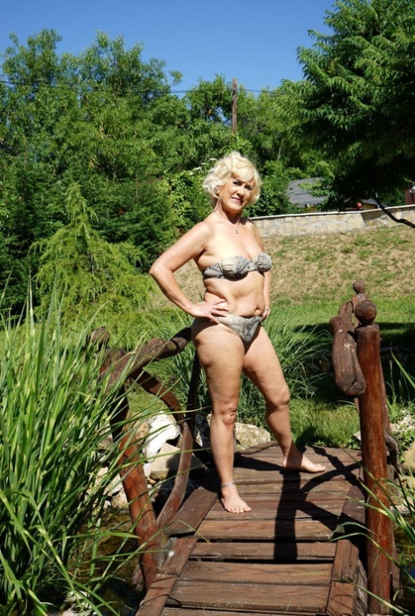 A mature woman with blonde hair removes her swimsuit while walking around her house.