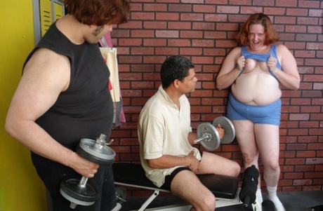 In the weight room, Adrienne, a fat and older redhead, engages in self-pleasure by sucking and fucking two cocks.