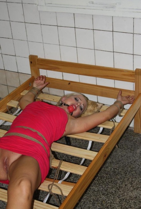 Ball gagged blond female struggles against rope restraints while wearing dress that has been raised.