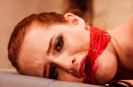 On a mattress, the redhead female is gagged and left face down while being tied up in hogtied positions.