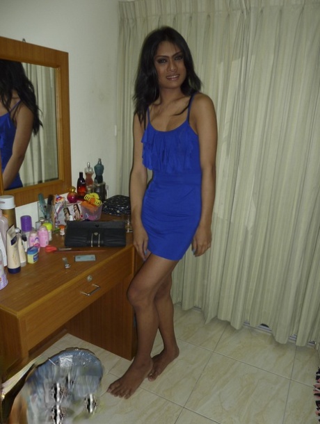 In his dark-skinned blue dress, a ladyboy with dark skin flashes black panties while in a single bedroom.