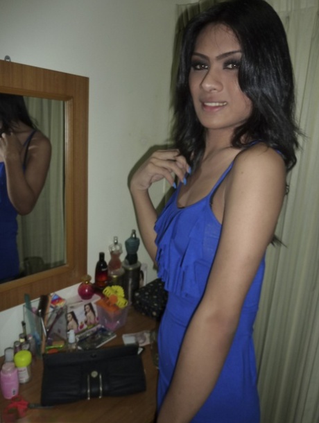 A ladyboy with dark skin and blue attire flashes black panties in his dark brown dress in the bedroom.