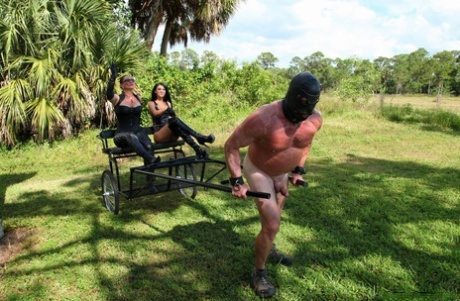 Femdoms Esmi Lee & Venus impart a lesson to their naked muscular slave outside of the enclosure.