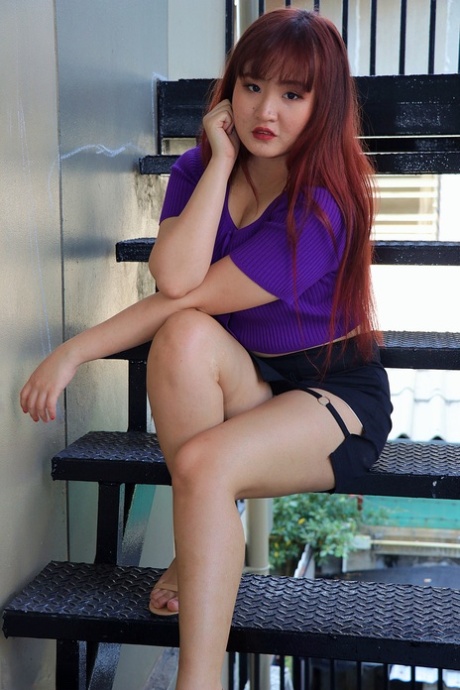 Redheaded Asian beauty, wearing her sexy skirt and purple top, poses on the stairs at one side.